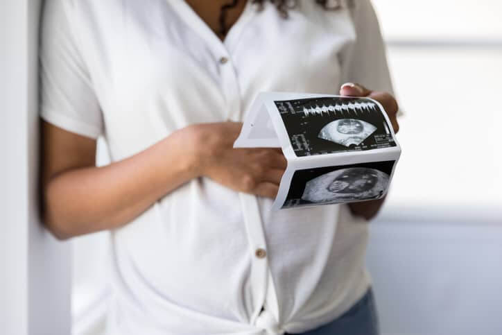 The unrecognizable pregnant woman looks at ultrasound photos of her baby while touching her belly.