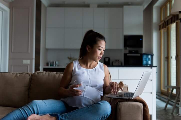 As she sits on her couch and has a glass of water, the mid adult pregnant woman uses her laptop to email.