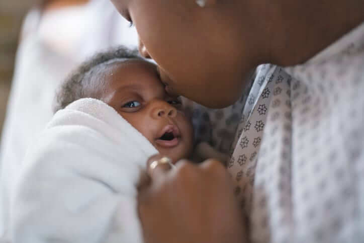 A young mother is kissing her newborn son on the forehead. They are at the hospital. The mother is wearing a hospital gown and the baby is wrapped in a white blanket.